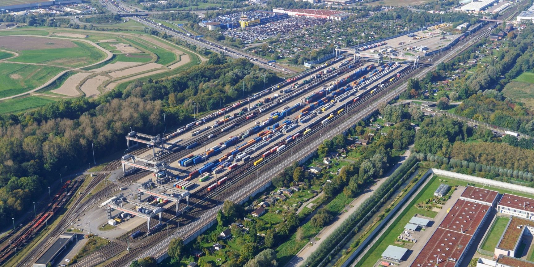 Eine Weitwinkel- oder Luftaufnahme des DUSS-Terminals Hamburg-Billwerder, die die massiven Portalkräne über zahlreichen Gleisen zeigt, auf denen Güterzüge mit Containern und Sattelaufliegern beladen werden. Die Szene vermittelt die enorme Größe der Logist
