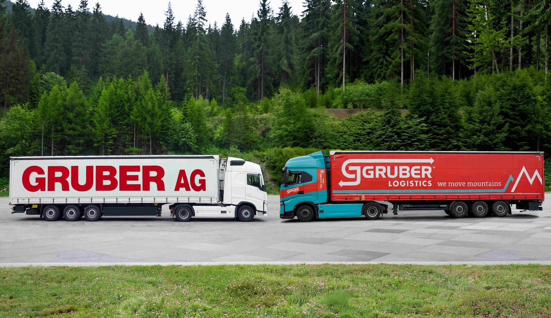 Lkw mit Trailer der Gruber AG und Lkw mit Trailer von GRUBER Logistics mit dem Slogan we move mountains stehen nebeneinander vor einem Nadelwald