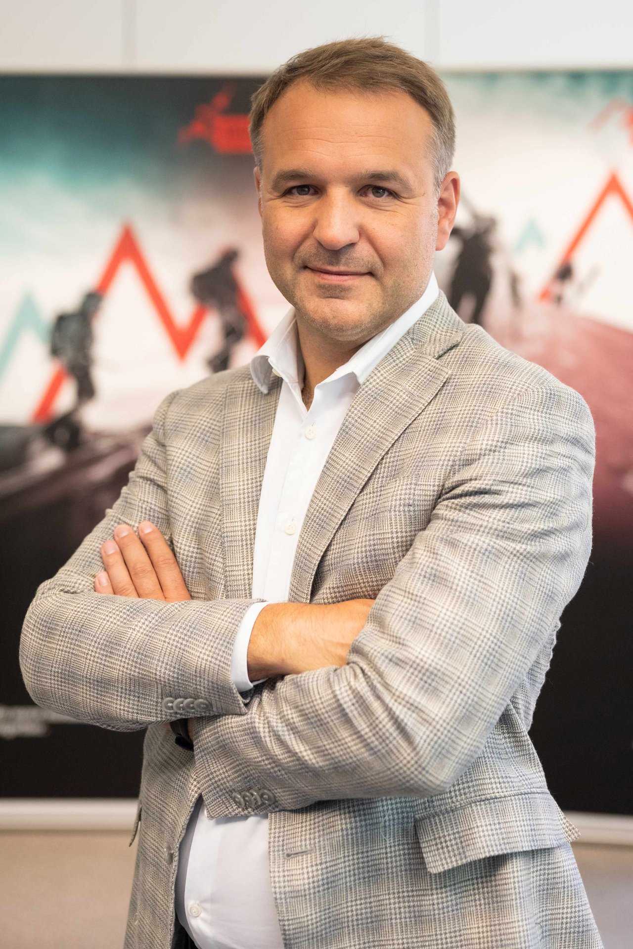 Porträt von Martin Gruber, CEO von GRUBER Logistics, mit verschränkten Armen in hellem karierten Sakko und weißem Hemd vor einer Bildwand mit dem Bergmotiv von GRUBER Logistics